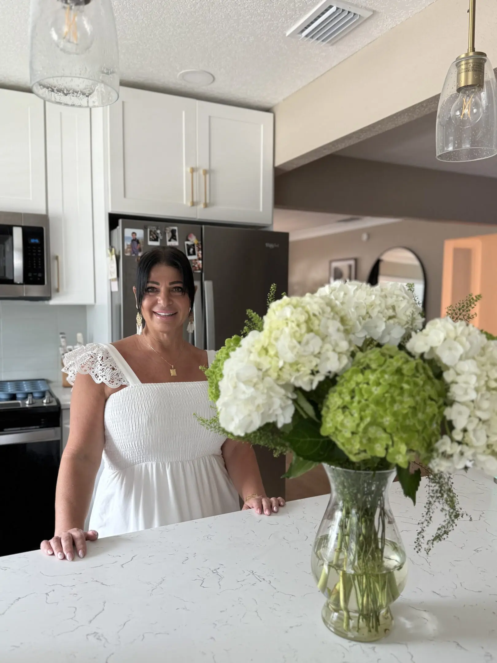 A woman in a white dress smiling in a modern kitchen with flowers.